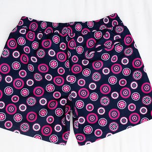 May include: Navy blue shorts with an all-over pattern of pink and white floral designs. The shorts have an elastic waistband and a relaxed fit. The fabric appears to be a lightweight material, suitable for warm weather. The shorts are laid flat on a white surface.