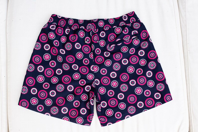 May include: Navy blue shorts with an all-over pattern of pink and white floral designs. The shorts have an elastic waistband and a relaxed fit. The fabric appears to be a lightweight material, suitable for warm weather. The shorts are laid flat on a white surface.