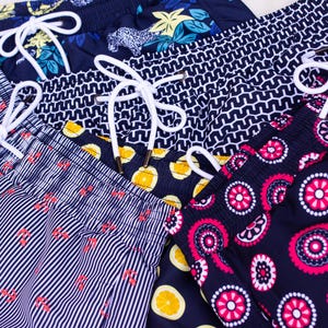 May include: Close-up of several pairs of swim trunks with various patterns. Designs include vertical stripes with red accents, geometric shapes, floral, and fruit patterns. Each pair has a white drawstring.