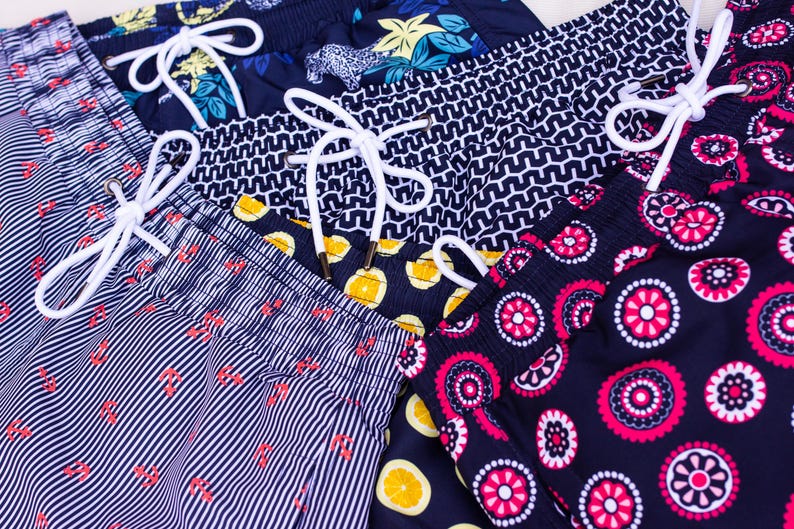 May include: Close-up of several pairs of swim trunks with various patterns. Designs include vertical stripes with red accents, geometric shapes, floral, and fruit patterns. Each pair has a white drawstring.