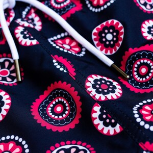 May include: Close-up of navy blue fabric with a repeating floral pattern in red, black, and white. A white drawstring with gold-colored aglets is visible. The design features stylized floral shapes.
