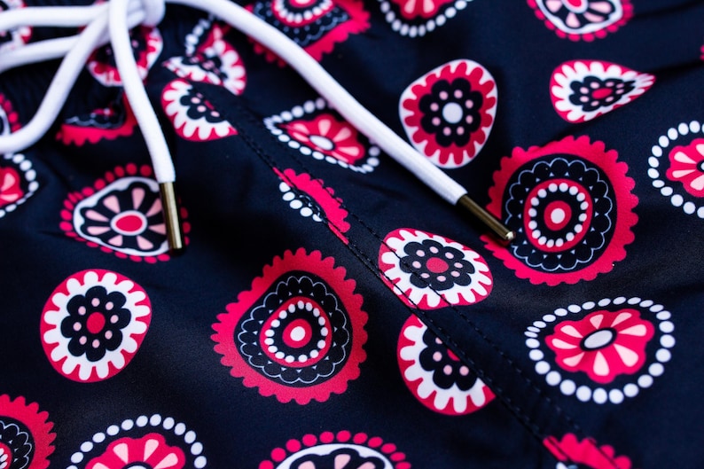 May include: Close-up of navy blue fabric with a repeating floral pattern in red, black, and white. A white drawstring with gold-colored aglets is visible. The design features stylized floral shapes.