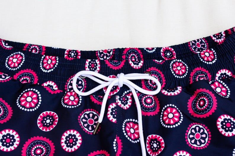 May include: Close-up of navy blue swim trunks with a repeating pattern of pink and white floral designs. The waistband is elasticized and features a white drawstring with gold-colored metal aglets.