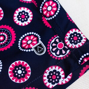 May include: Close-up of a navy blue fabric with a repeating floral pattern in pink and white. The design features stylized flowers and circular shapes. A small, white label with a black triangle is visible.