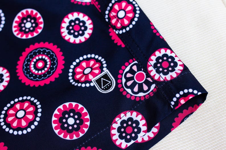 May include: Close-up of a navy blue fabric with a repeating floral pattern in pink and white. The design features stylized flowers and circular shapes. A small, white label with a black triangle is visible.