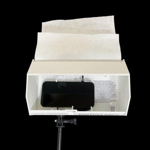 May include: A white Cool Fence Cam housing unit mounted on a black tripod stand. The device features a rectangular opening containing a black smartphone and a printed instruction label. A white paper sheet is draped over the top of the enclosure.
