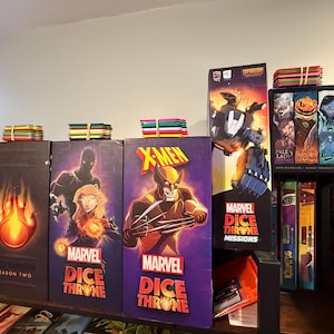 May include: A collection of Marvel Dice Throne board game boxes arranged on a shelf, featuring character artwork of Wolverine, Black Panther, and Captain Marvel, alongside a Dice Throne Missions box and stacks of colorful 3.5-inch floppy disks.