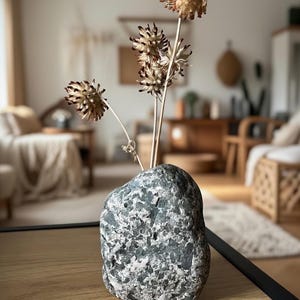 May include: A decorative vase made from a gray and white speckled stone, holding dried beige flowers with brown tips. The vase sits on a light wood surface, with a blurred background of a living room.
