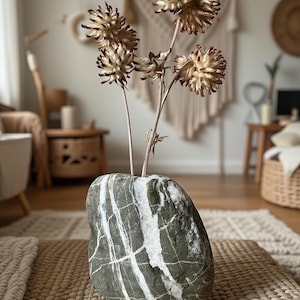 Puede incluir: Un jarrón de piedra natural con vetas blancas, que contiene flores secas de color beige y marrón. El jarrón está sobre una estera tejida, con un interior de estilo bohemio de tonos neutros en el fondo. El jarrón mide unos 20 cm de alto.