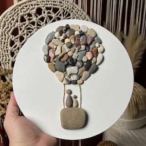 Könnte beinhalten: Runde, weiße Wanddekoration mit einem Heißluftballon-Design. Der Ballon besteht aus einer Vielzahl farbiger Kieselsteine, mit einem kleinen Korb darunter, der zwei Kieselsteinfiguren enthält. Ein einzigartiges, handgefertigtes Kunstwerk.