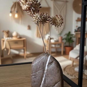 Peut inclure: Un vase en pierre gris foncé, lisse et arrondi, présentant une veine minérale blanche verticale proéminente au centre. Trois têtes de fleurs séchées, épineuses et brun clair, sur des tiges fines, sont disposées dans l'ouverture.