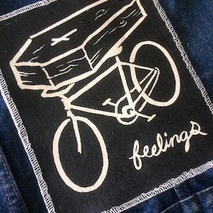 May include: Black patch with a white coffin on a bicycle, with the word "feelings" in cursive. The patch is sewn onto a blue denim jacket.