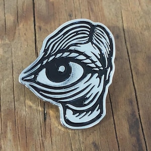 May include: A silver-colored pin with a black and white eye design. The pin features a detailed illustration of an eye with eyelashes and a stylized eyebrow. The eye is set against a silver background with a black outline.