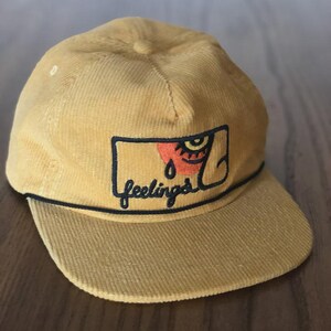 Könnte beinhalten: Senfgelbe Cord-Baseballkappe mit gebogenem Schirm. Die Kappe hat einen rechteckigen Aufnäher mit einer Augen- und Tränen-Grafik und dem Wort "feelings" in schwarzer Schrift.
