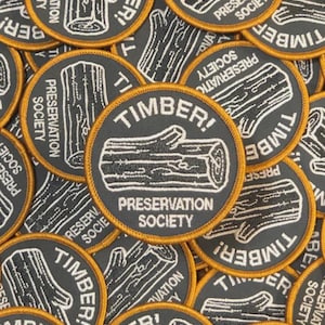 May include: A collection of circular patches with a gray background and a gold border. Each patch features a white illustration of a log and the text "TIMBER!" and "PRESERVATION SOCIETY".