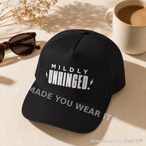 Op de afbeelding: Een zwarte baseballpet met de witte tekst MILDLY UNHINGED en kleine bliksemschichtpictogrammen aan weerszijden. De pet heeft een gestructureerd ontwerp met vijf panelen, een gebogen klep en is volledig gemaakt van zwarte stof.