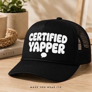 Peut inclure: Une casquette de type trucker noire avec un panneau arrière en filet et un panneau avant solide. L'avant présente le texte blanc "CERTIFIED YAPPER" dans une police arrondie et grasse, avec une petite icône de bulle de dialogue blanche.