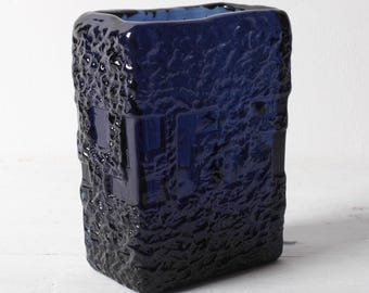 May include: A rectangular cobalt blue glass vase featuring a thick, textured exterior surface with a rough, craggy appearance. The glass is translucent, allowing light to pass through the deep blue material, and the top opening is wide and smooth.