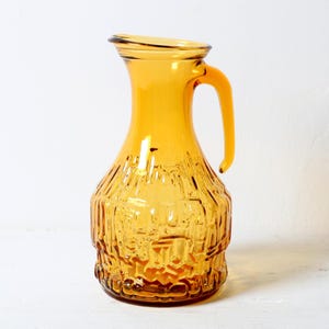 Op de afbeelding: Een karaf van doorschijnend amberkleurig glas met een brede, ronde basis en een gestructureerd, geometrisch reliëfpatroon. De karaf heeft een gladde, uitlopende hals, een gebogen schenktuit en een stevig, geïntegreerd handvat aan de zijkant.