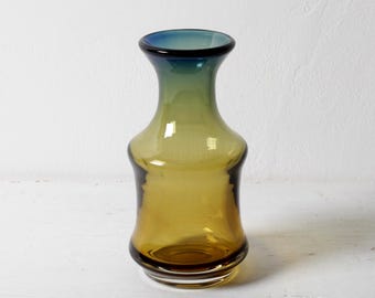 May include: A glass vase featuring a gradient colour transition from a deep amber base to a lighter olive green midsection, culminating in a dark blue rim. The vessel has a wide, rounded bottom that tapers into a slender neck with a flared opening.