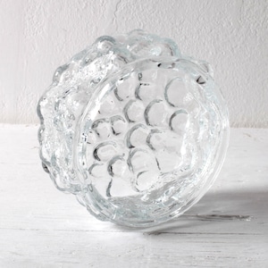 Puede incluir: Un cuenco de vidrio transparente con un patrón texturizado en forma de burbujas. El cuenco tiene una forma redondeada con un borde ondulado y texturizado. El vidrio parece grueso y pesado, reflejando la luz. El cuenco es probablemente para fines decorativos o de servicio.