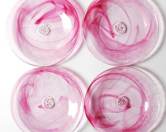 Op de afbeelding: Een set van vier ronde glazen borden met een wervelend magenta en roze gemarmerd patroon in het heldere glas. Elk bord heeft een cirkelvormig, getextureerd medaillon in het midden en een klein reliëflogo bij de bovenrand van het bord.