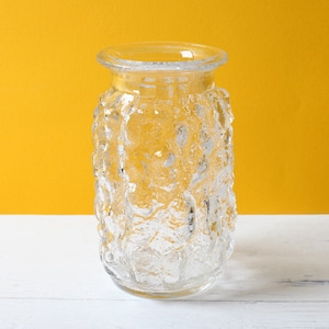 Puede incluir: Un jarrón de vidrio transparente con una superficie texturizada y burbujeante. El jarrón tiene una abertura ancha y un borde ligeramente ensanchado. El fondo es de color amarillo liso y el jarrón está sobre una superficie blanca.