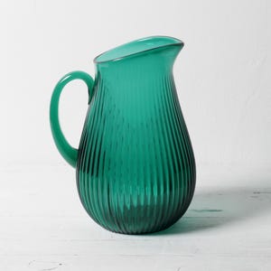 Può includere: Una brocca in vetro verde smeraldo traslucido caratterizzata da un corpo arrotondato con dettagli a coste verticali e un manico curvo. Il recipiente presenta un beccuccio ampio e aperto sulla parte superiore e una finitura liscia.