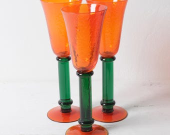Op de afbeelding: Drie hoge drinkglazen met getextureerde oranje kelken, slanke groene stelen en brede oranje ronde voeten. De glasset vertoont een levendig contrast tussen de feloranje bovendelen en de donkergroene stelen en de voeten van de glazen.