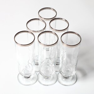 Puede incluir: Un juego de seis vasos transparentes con forma cilíndrica alta y base redondeada. Cada vaso presenta un borde metálico plateado liso en la parte superior. El material de vidrio es transparente y liso, diseñado para servir bebidas.
