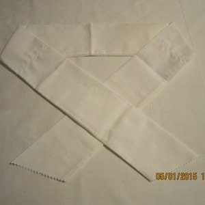 May include: A white fabric sash with a zig-zag edge. The sash is folded in half and laid flat on a white surface.