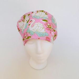 May include: A pink fabric cap featuring a swan and floral pattern. The swans are white with orange beaks and are set against a backdrop of light blue water and green foliage. The flowers are pink and white.