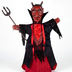 Puede incluir: Una marioneta de diablo con cara roja, cuernos y orejas puntiagudas. Viste un abrigo negro con ribetes rojos, una corbata de satén roja y una falda roja. La marioneta sostiene un tridente gris oscuro en su mano derecha y tiene brazos.