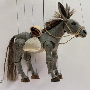 Puede incluir: Títere de burro de madera gris con larga melena y cola marrón, y brida de cuerda. Tiene un saco pequeño y una manta enrollada en la espalda. Las patas del burro son articuladas y tiene pezuñas de madera. El texto "c. 36 44cm" está en la esquina inferior izquierda.