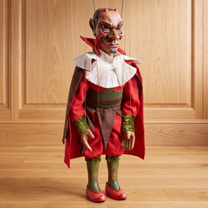 May include: A marionette puppet featuring a red-faced character with small horns, wearing a red and brown outfit with a white pleated collar and a red cape. The figure has green accents on the sleeves and legs, and red shoes, suspended by thin control strings.