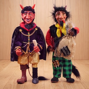 May include: Two devil marionettes with strings attached. The left puppet wears a purple cape, gold pants, and one brown boot. The right puppet has messy black hair, a yellow scarf, green and black plaid pants, and carries a burlap sack with a chain.