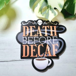 May include: A black and orange sticker with the text "DEATH BEFORE DECAF" and a small skull graphic. The sticker features a coffee cup graphic at the bottom. The sticker is on a white surface.