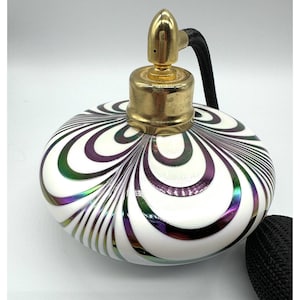 Può includere: Un flacone di profumo in vetro bianco con un tappo dorato e un ugello spray nero. Il flacone presenta un design a spirale iridescente in tonalità di viola, verde e oro.