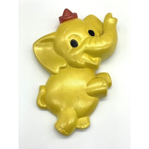 May include: A yellow plastic elephant figurine with a red hat. The elephant is smiling and has large black eyes. It is a vintage toy.