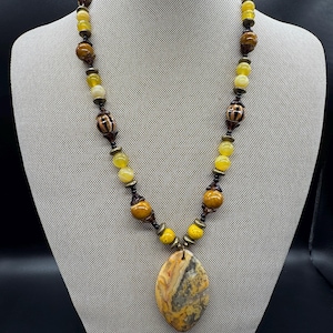 May include: A beaded necklace with a large, oval pendant. The necklace features yellow, brown, and tan beads, with decorative metal accents. The pendant is a mix of tan, gray, and orange colors.