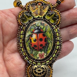 Op de afbeelding: Handgemaakte hangerketting met een ovale afbeelding van een lieveheersbeestje. De hanger is omgeven door ingewikkeld kralenwerk in goud, groen en bruin, met vlinder- en bladmotieven. Uniek sieraad.