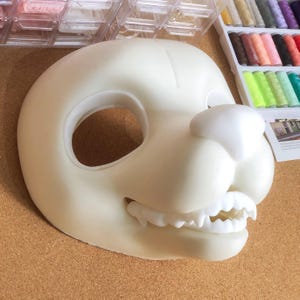 May include: A white, unpainted, resin fursuit base for a canine character. The base has a large eye hole, a nose, and a mouth with teeth.