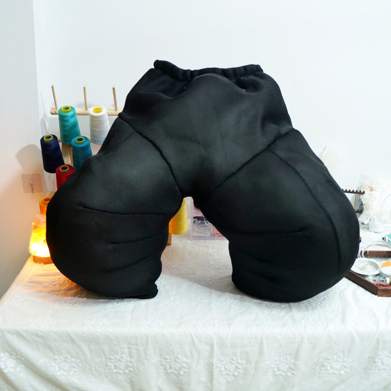 May include: Black, padded, U-shaped pillow designed for sitting or lounging. The pillow has a mesh fabric exterior and a zipper closure.