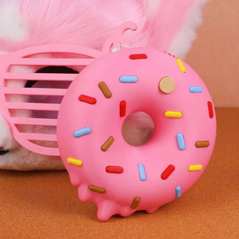 May include: A pink donut-shaped stress ball with colourful sprinkles. The donut has a hole in the centre and a melted frosting effect on the bottom. A pink, plastic, shutter-style sunglasses is in the background.