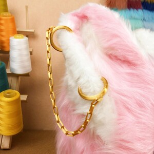 May include: A gold-colored chain is attached to a fluffy, pink and white faux fur item. The chain connects to two gold-colored crescent-shaped clasps. Spools of thread in various colors are visible in the background.