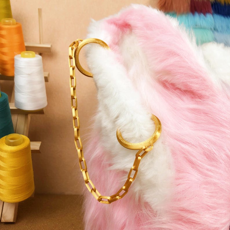 May include: A gold-colored chain is attached to a fluffy, pink and white faux fur item. The chain connects to two gold-colored crescent-shaped clasps. Spools of thread in various colors are visible in the background.