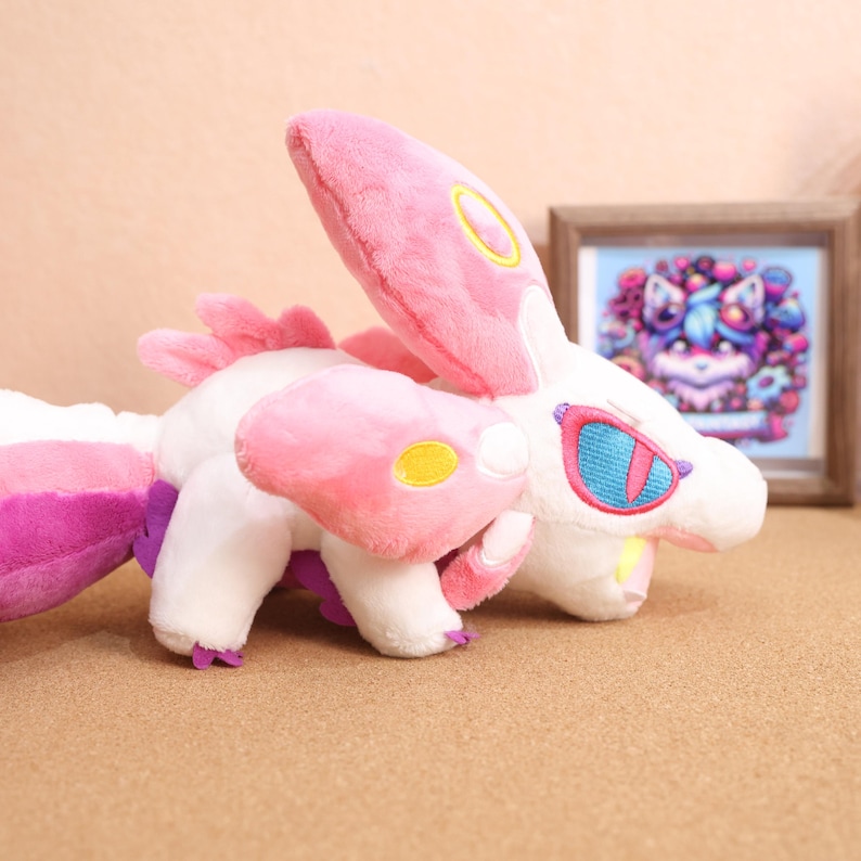 May include: A pink and white plush toy with a long tail and large ears. The toy has yellow and blue accents on its face and body.