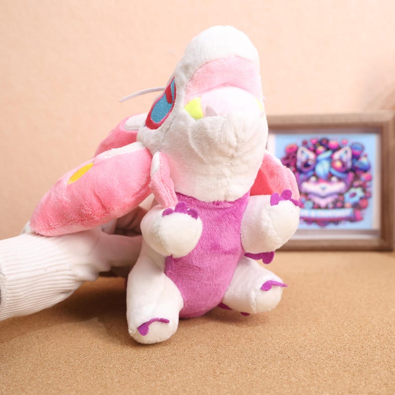 May include: A pink and white plush toy with a large pink wing on one side. The toy has a white face with green eyes and a pink mouth.
