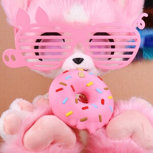 May include: A pink plush character wearing pink shutter shades and holding a pink donut with colourful sprinkles. The character has pink fur and paws. The background is a light brown colour.