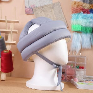 May include: A light gray baby safety helmet on a white mannequin head. The helmet is padded and has a chin strap with a bow. The background includes sewing supplies and fabric samples.